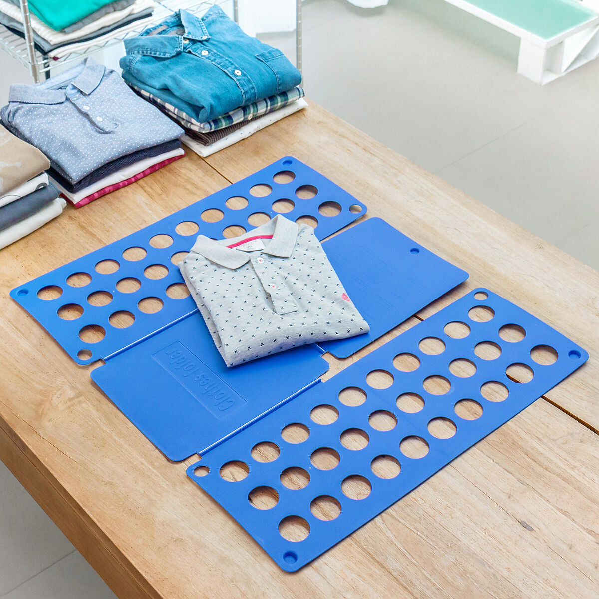 Blaues Wäschefaltbrett auf einem Tisch mit gefaltetem Hemd und weiteren Kleidungsstücken