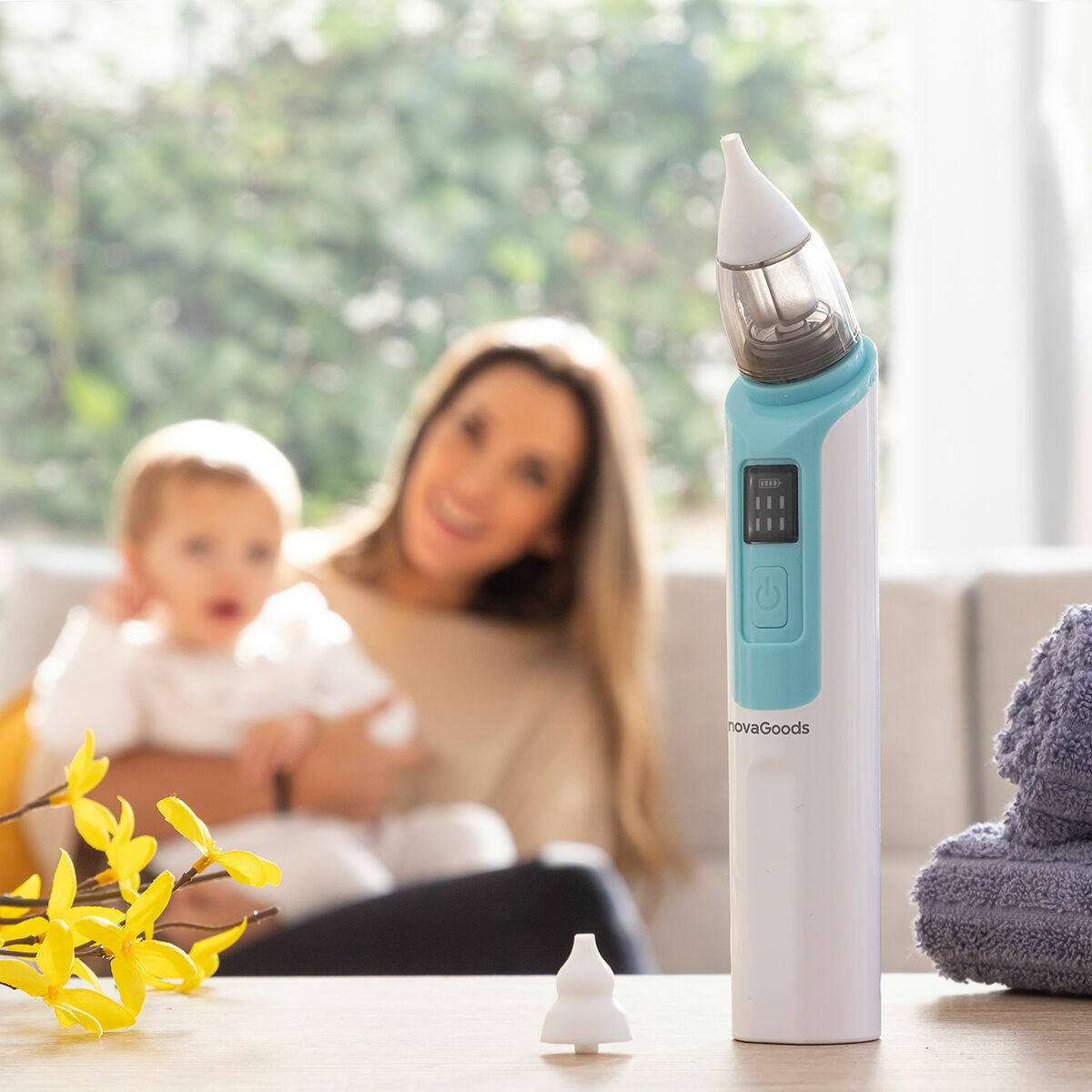 Weiß-blauer Nizi-Nasensauger auf einem Tisch mit zweitem Silikonaufsatz, Mutter und Kind im Hintergrund